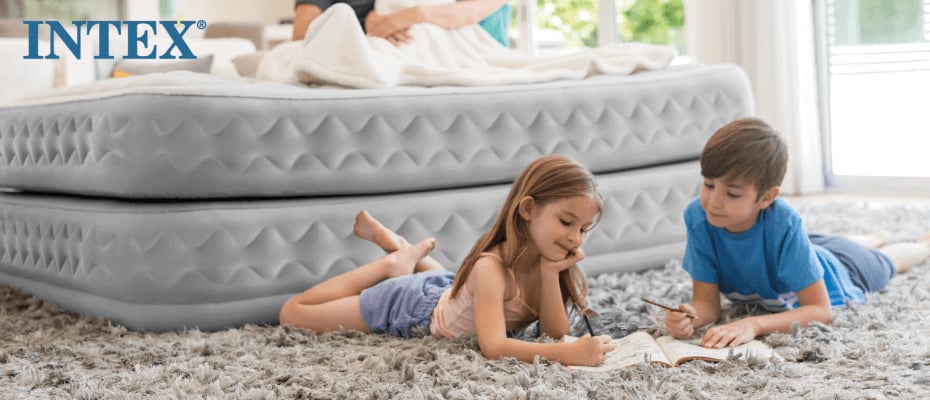 Das Bild zeigt zwei Kinder, die auf einem Teppich vor einem grauen, aufblasbaren Intex-Luftbett liegen. Sie sind mit Zeichnen oder Schreiben beschäftigt. Im Hintergrund ist das Luftbett in einem hellen, gemütlichen Raum zu sehen, der eine entspannte Atmosphäre vermittelt.