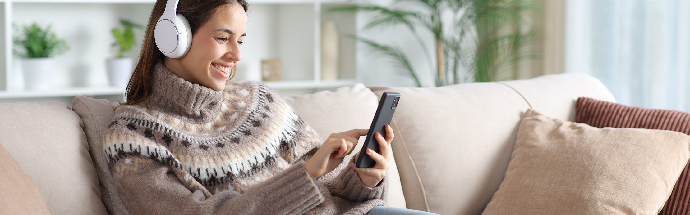 Eine lächelnde junge Frau mit weißen Over-Ear-Kopfhörern entspannt auf einem hellen Sofa und bedient ihr Smartphone, während sie Musik oder einen Podcast hört.