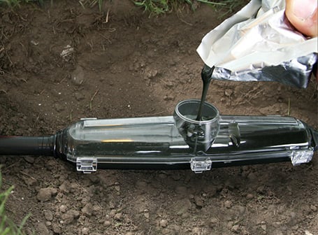 In einem Erdloch wird dunkles Gießharz aus einem Beutel in eine transparente Kunststoffmuffe gefüllt, um eine elektrische Kabelverbindung im Boden zu isolieren und abzudichten.