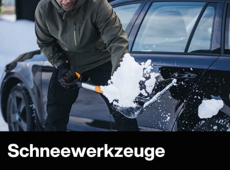 Eine Person befreit mit einer Schneeschaufel von Fiskars ein schwarz glänzendes Auto in einer verschneiten Umgebung von Schneemassen.