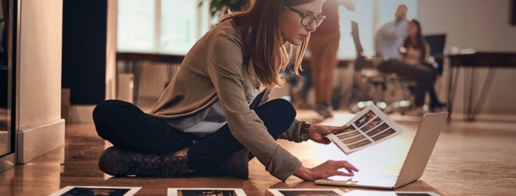 Eine junge Frau mit Brille sitzt mit gekreuzten Beinen auf einem Holzboden und betrachtet Ausdrucke vor einem geöffneten Laptop, während im Hintergrund verschwommene Personen zu sehen sind.