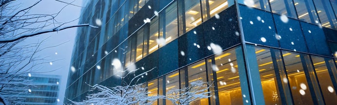 Glasfassade eines modernen Bürogebäudes bei Schneefall mit beleuchteten Innenräumen.