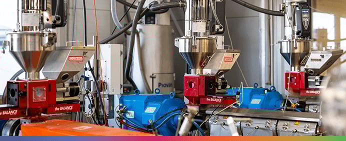 Eine Innenansicht einer Industrieanlage mit mehreren silbernen Trichtermaschinen, blauen Motoren und komplexen Rohrleitungssystemen zur professionellen Herstellung von Filamenten.