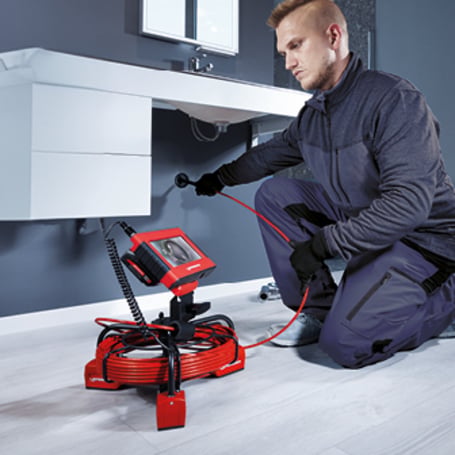 Ein Handwerker führt ein rotes Kamerakabel zur Rohrinspektion in einen Wandanschluss unter einem Waschbecken ein, während das Bild auf einem Monitor übertragen wird.