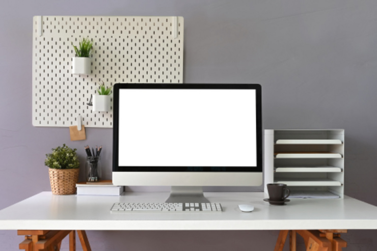 Schreibtisch mit Computer, Pflanzen, Stifte, Kaffeetasse und Organizer vor einer grauen Wand.