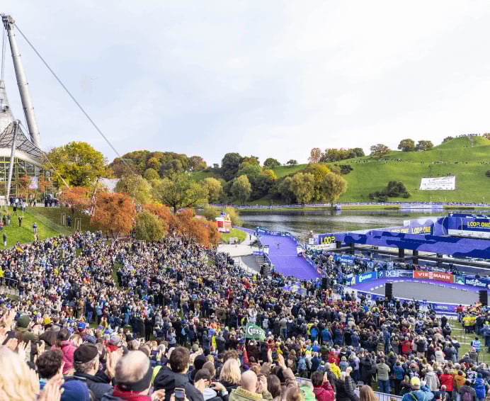 Eine große Menschenmenge bejubelt von einem Grashang aus ein Sommer-Biathlon-Rennen im Münchener Olympiapark, bei dem die Strecke an einem See und unter der markanten Zeltdachkonstruktion verläuft.