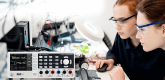 Zwei konzentriert arbeitende junge Frauen mit Schutzbrillen betrachten einen Monitor in einem Elektroniklabor, während im Vordergrund ein digitales Multimeter von Rohde & Schwarz präzise Messwerte anzeigt.