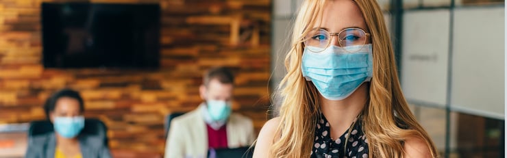 Eine Frau mit Brille und medizinischer Maske steht im Vordergrund eines Büros, während zwei weitere maskierte Personen im Hintergrund sitzen.