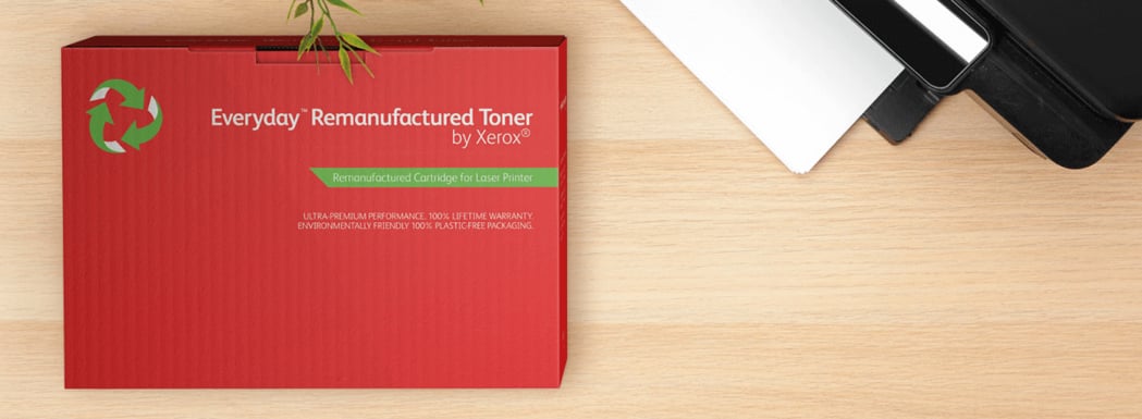 Dieses Bild zeigt einen roten Karton mit der Aufschrift "Everyday Remanufactured Toner by Xerox" in dem Xerox Toner enthalten ist. Der Karton liegt auf einer hellen Holzoberfläche neben einem diagonal angeordneten schwarzen Drucker, der im Anschnitt zu sehen ist.