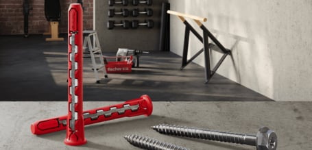  Zwei große, rot-metallische Dübel, die in einem 90-Grad-Winkel voreinander auf einem Betonboden liegen, mit zwei dazugehörigen Schrauben im Vordergrund und Fitnessgeräten, wie Hanteln und Turnstangen, im Hintergrund.