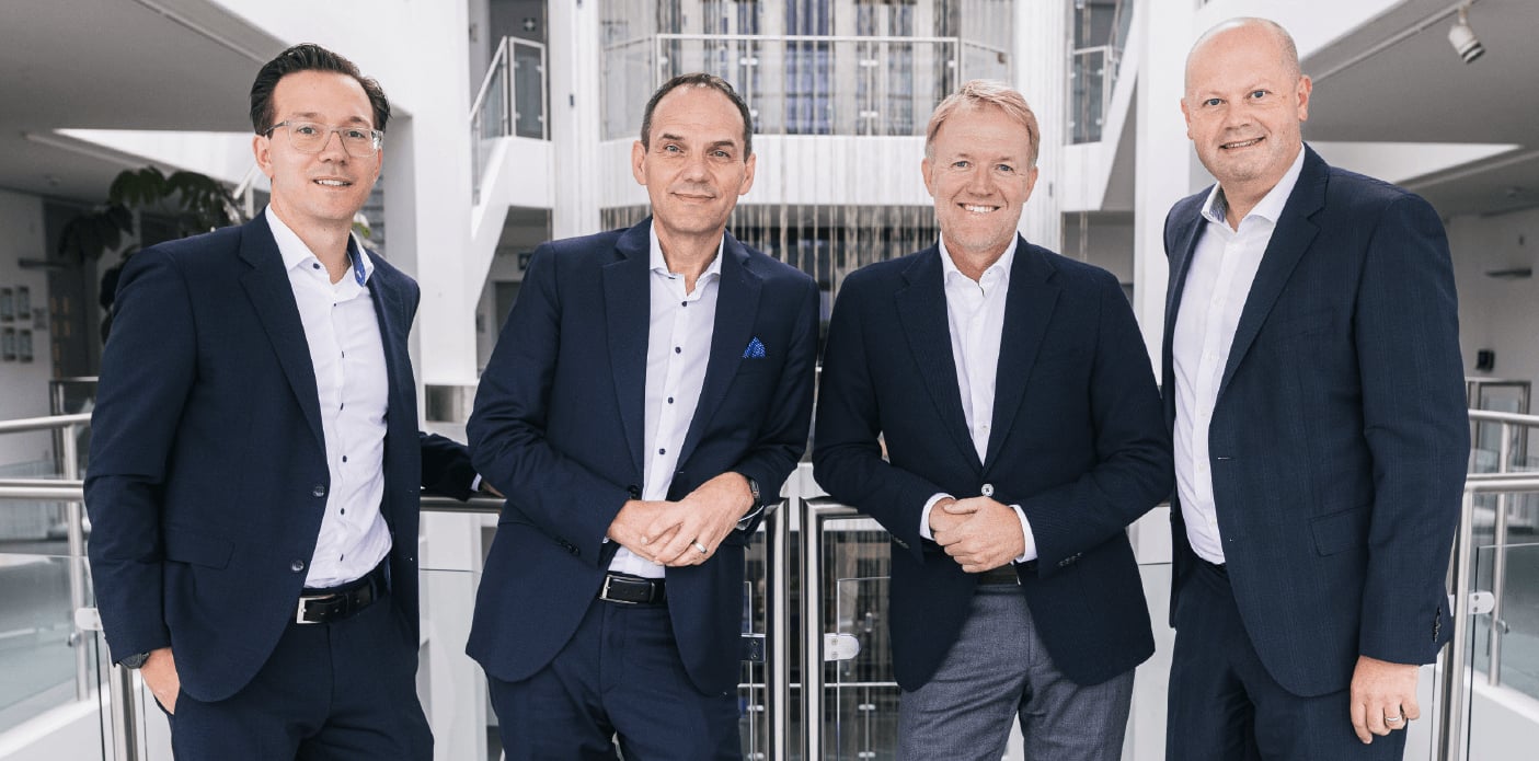 Klarer Kundenfokus – die Geschäftsführung von Conrad Electronic: Dr. Sebastian Dehnen, Ralf Bühler, Dr. Daniel Saga und Guy Dann (v.l.n.r.) in dunklen Business-Anzügen und hellen Hemden stehen nebeneinander im Treppenhaus des Firmensitzes in Hirschau..