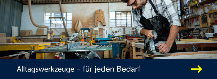 Person mit Gehörschutz und Schutzbrille bearbeitet in einer Werkstatt eine Holzplatte mit einer Handkreissäge.