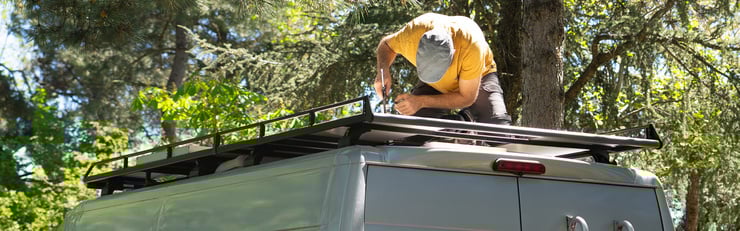 Eine Person montiert im Freien einen Dachträger auf dem Dach eines Fahrzeugs, umgeben von Bäumen.