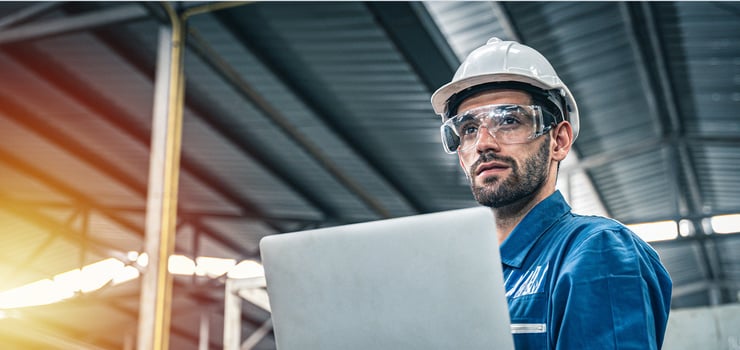 Ein Arbeiter mit weissem Schutzhelm in einer Halle mit einem Laptop in der Hand und Schutzbrillen auf.