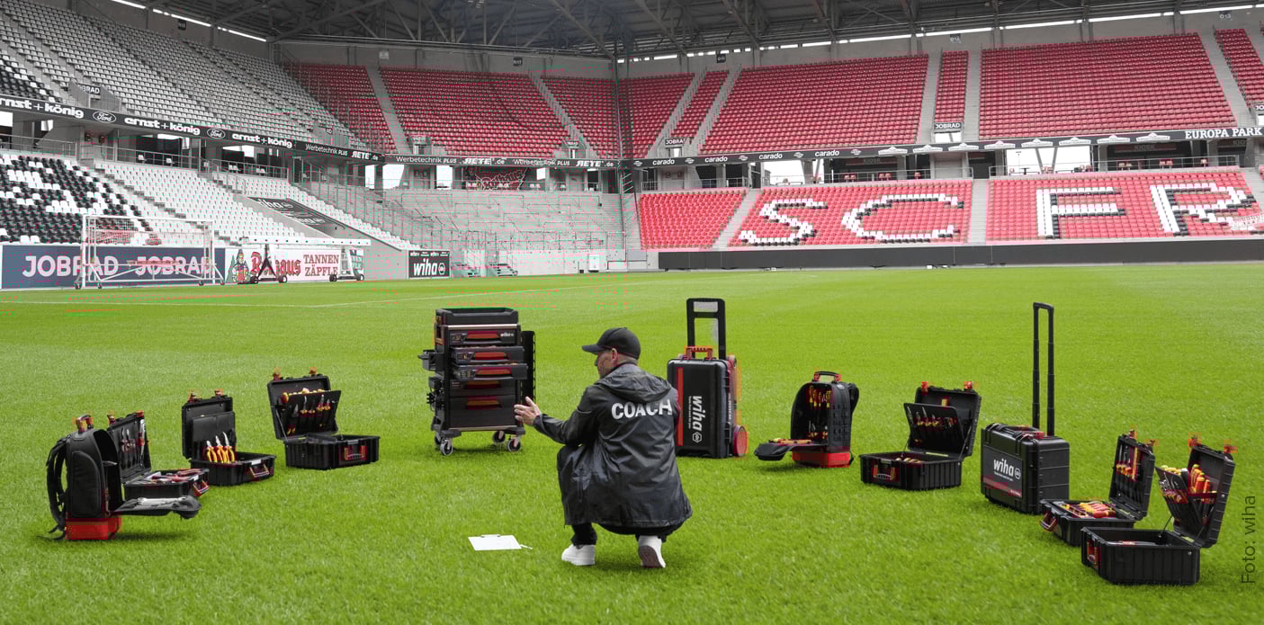 Das Bild zeigt eine Person im Sportstadion die sich einem Arrangement von Wiha Werkzeugkoffern zuwendet, die auf der Rasenfläche verteilt stehen.
