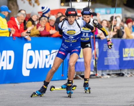 Zwei Biathletinnen auf Rollski in voller sportlicher Ausrüstung fahren während eines Sommerbiathlon-Wettkampfs an einer jubelnden Zuschauermenge vorbei.