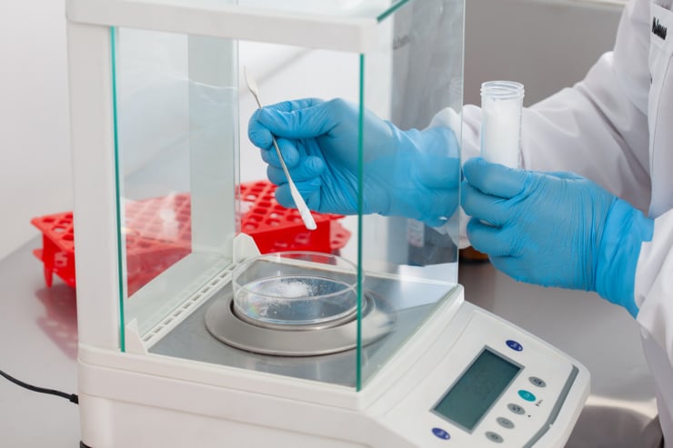 Wissenschaftler/in mit blauen Handschuhen wiegt eine Substanz in einer Laborschale unter einer Abzugshaube.