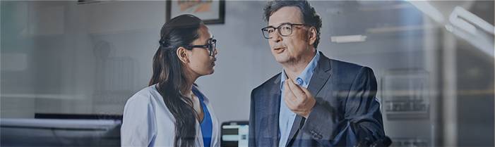 Frau mit dunklen Haaren, Mann mit Brille in Gespräch vertieft, Bürohintergrund. Zur ProcurePlus