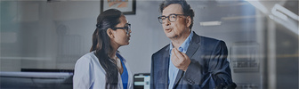 Woman with dark hair, man with glasses engrossed in conversation, office background. To ProcurePlus