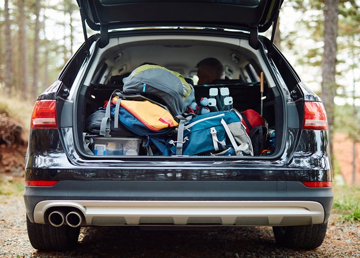 Ein Auto mit geöffnetem Kofferraum, vollgepackt mit Rucksäcken und Outdoor-Ausrüstung in einem Wald.