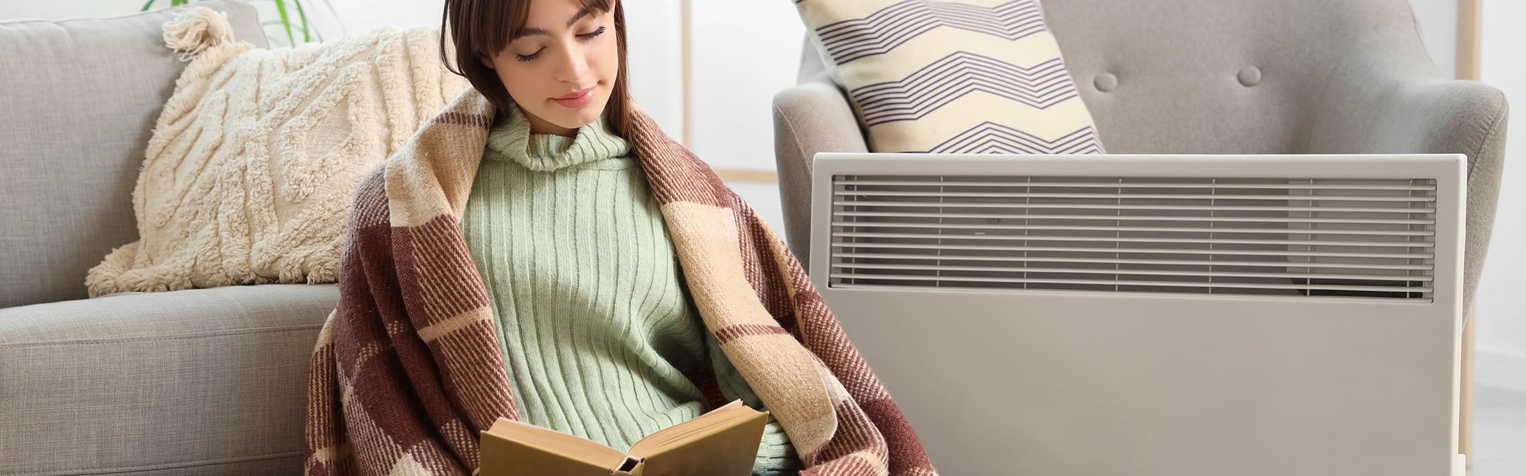 Une femme lit tranquillement un livre à côté d’un radiateur électrique dans un salon confortablement aménagé.