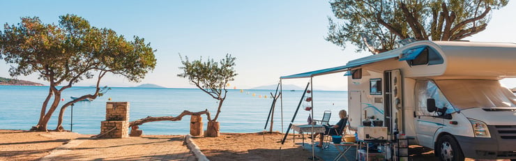 Un camping-car est garé sur un terrain de camping au bord de la mer, équipé d'un auvent et de chaises, avec vue sur mer.
