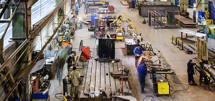 Grand hangar industriel avec plusieurs ouvriers travaillant sur des établis et des machines pour usiner et souder des pièces métalliques.