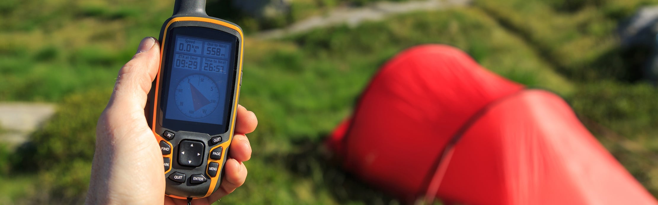 Une personne tient un GPS à la main, tandis qu'à l'arrière-plan, on aperçoit une tente dans un cadre naturel.