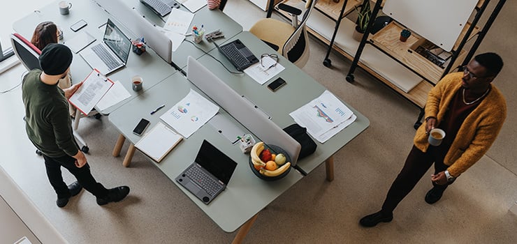 Plusieurs personnes travaillent dans un bureau moderne, à des bureaux communs, avec des ordinateurs portables, des documents et des tasses de café.