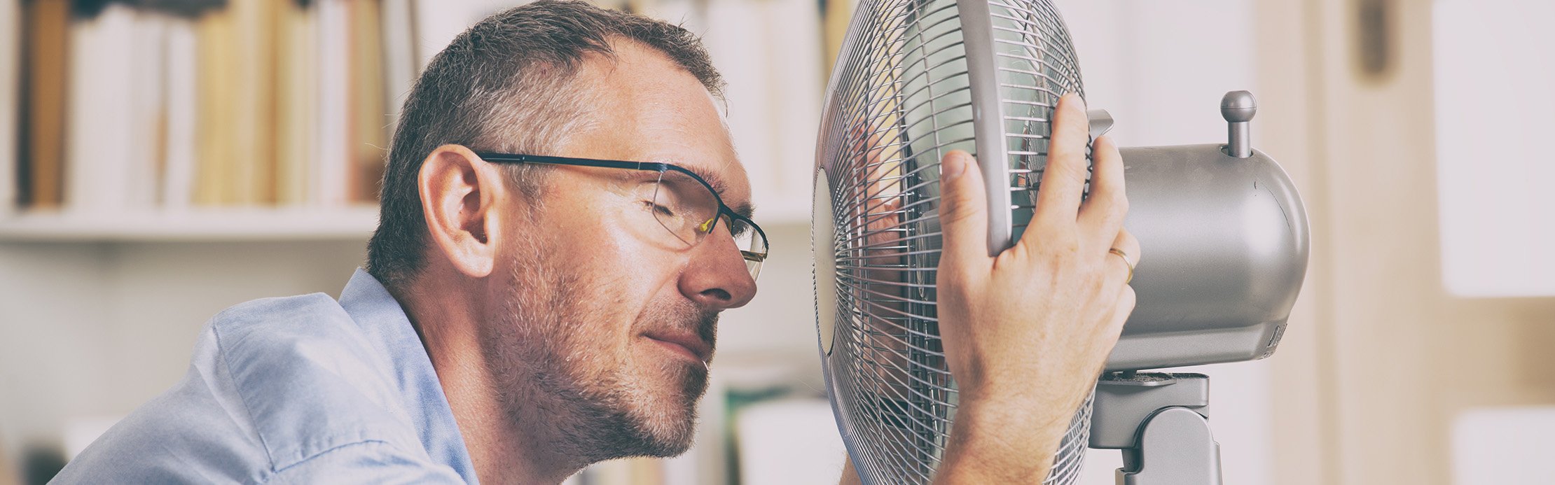 Un homme avec des lunettes est assis à son bureau et rapproche son visage d’un ventilateur pour se rafraîchir – Se rafraîchir en été