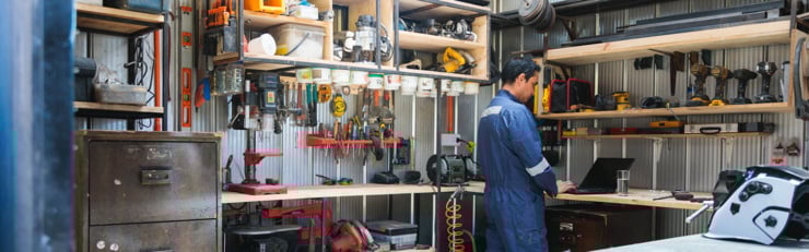 Un homme travaille sur un ordinateur portable dans un atelier bien équipé, entouré d’outils et de machines.