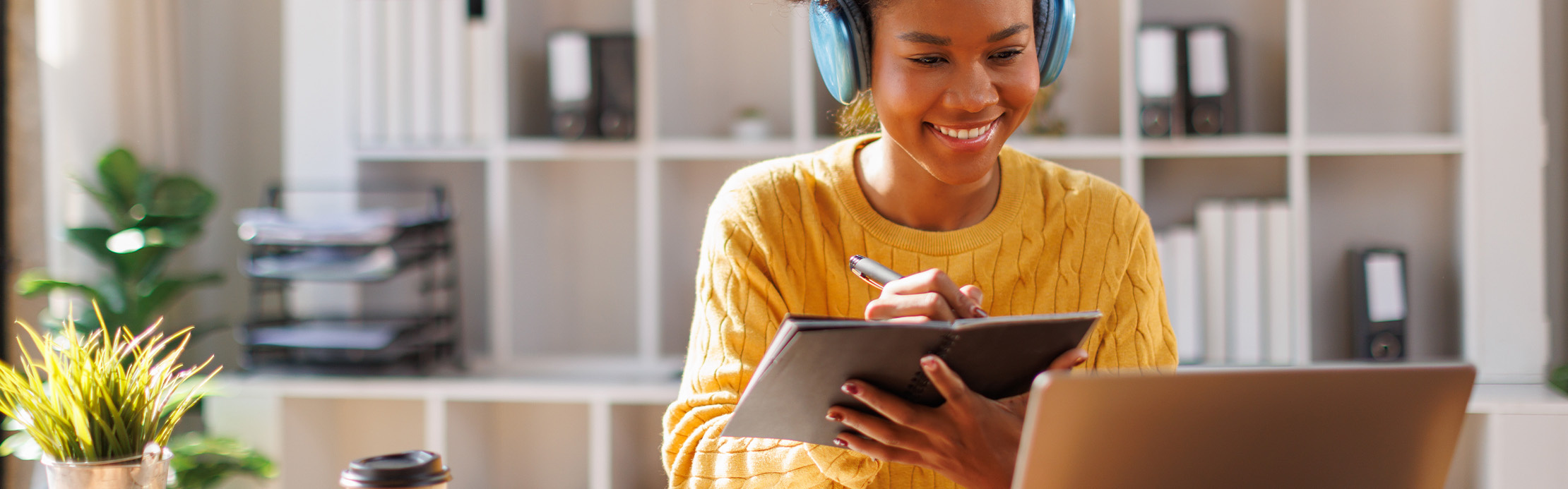 Une jeune femme portant un casque audio bleu est assise devant son ordinateur portable, en souriant, et prend des notes à la main dans un cahier.