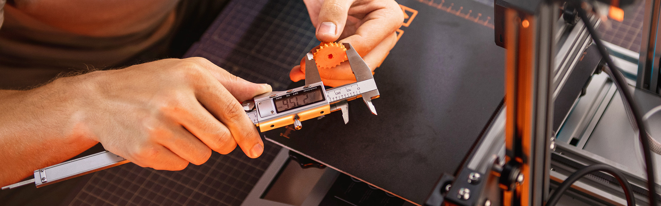 Une personne mesure un engrenage orange imprimé en 3D à l'aide d'un pied à coulisse numérique, à côté d'une imprimante 3D.