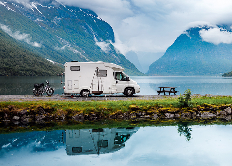 Un camping-car est garé au bord d’un lac paisible entouré de hautes montagnes avec reflet dans l’eau.