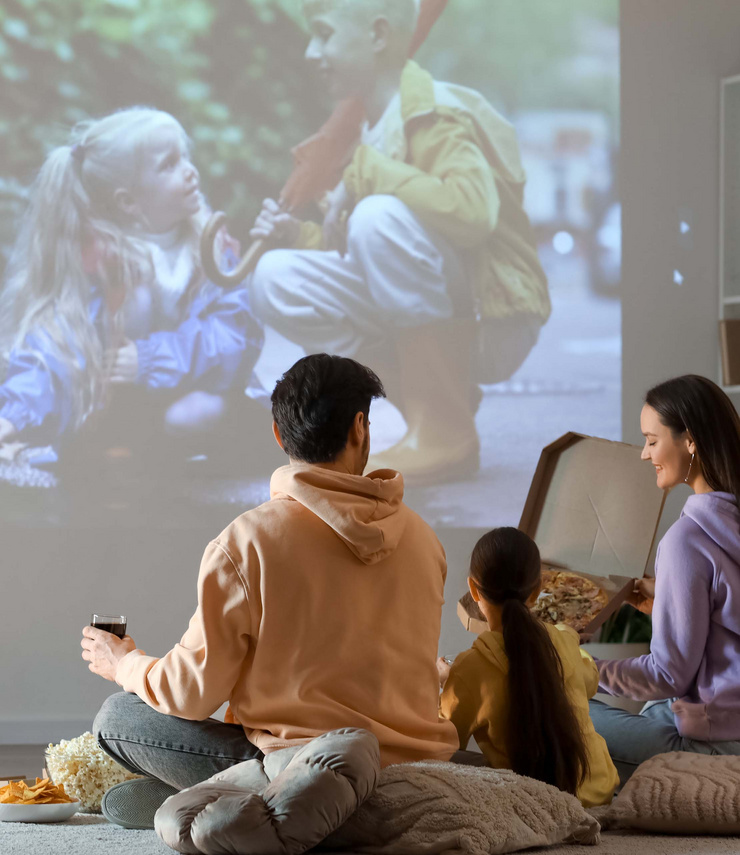 Une famille est assise confortablement sur des coussins, mange de la pizza et du pop-corn et regarde un film projeté sur le mur.