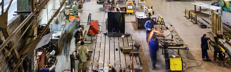 Grand hall industriel avec plusieurs personnes au travail, des machines, des établis et des opérations d'usinage des métaux en cours de production.