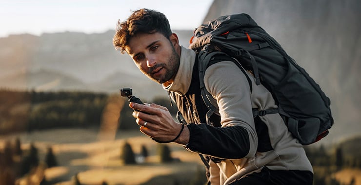 Un randonneur muni d'un sac à dos tient une caméra d'action à la main et se filme dans un paysage de montagne.