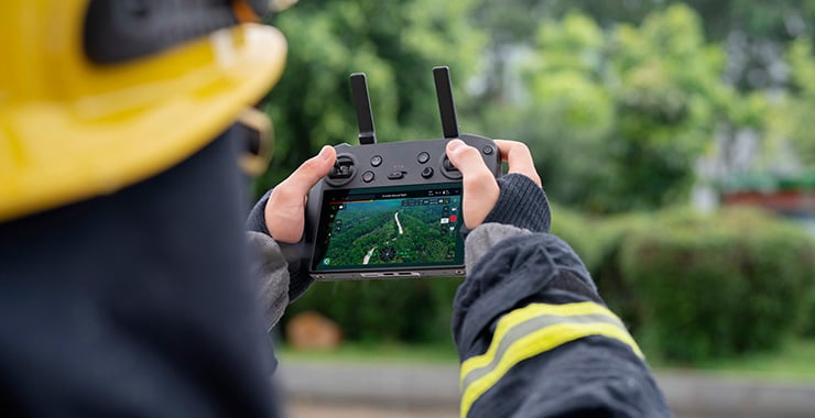 Une personne portant un casque de protection pilote un drone à l'aide d'une télécommande dotée d'un écran affichant des images en direct, à l'extérieur.