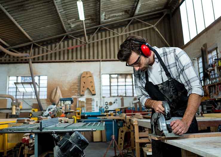 Une personne portant des lunettes de protection et des protections auditives découpe une plaque de bois à l'aide d'une scie circulaire à main dans un atelier.