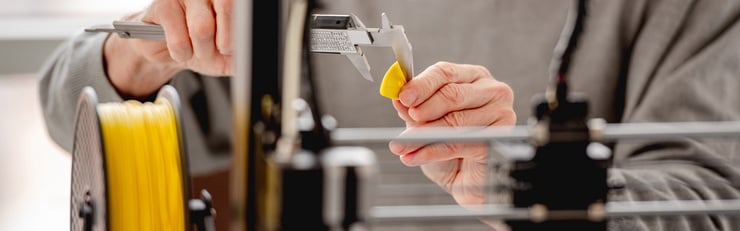 Une personne mesure avec un pied à coulisse une petite pièce jaune imprimée en 3D à côté d’une bobine de filament jaune sur une imprimante 3D.