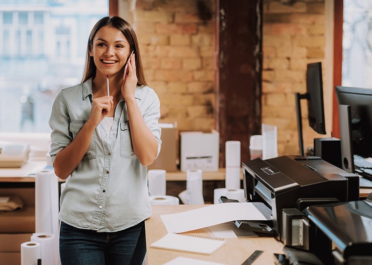 Une personne téléphone dans un bureau ou une salle d'impression, debout à côté d'un bureau équipé d'une imprimante, de rouleaux de papier et d'appareils de bureau.