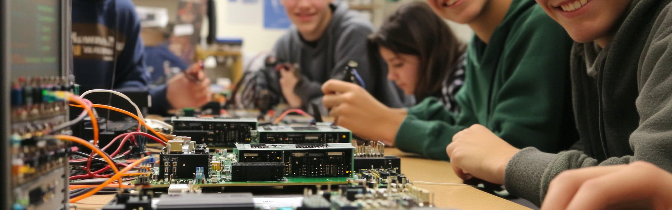 Des élèves travaillent sur une table avec des circuits imprimés et des câbles pendant un cours de technologie. Un adolescent sourit à la caméra.