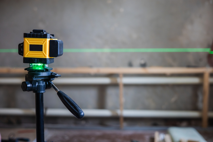 Un niveau laser jaune et noir monté sur un trépied projette une ligne laser verte horizontale sur un chantier, avec en arrière-plan un mur en béton et un échafaudage en bois