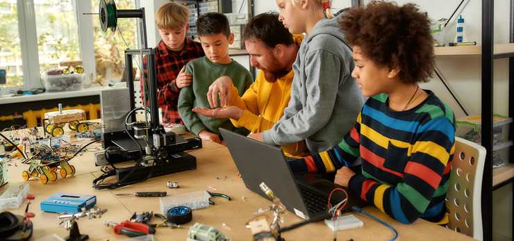 Un enseignant explique le fonctionnement des composants électroniques à un groupe de quatre enfants assis à une table équipée d'une imprimante 3D et d'un ordinateur portable.