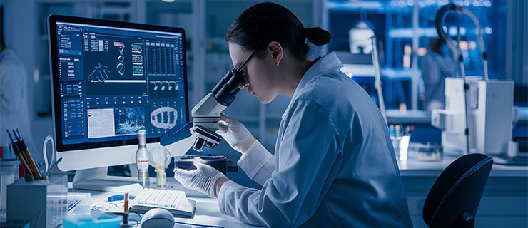 Une scientifique de laboratoire observe un échantillon au microscope, tandis qu’une analyse d’ADN s’affiche sur l’écran situé derrière elle. 