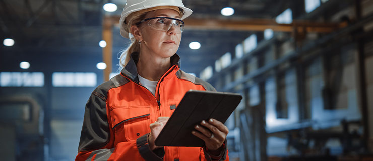 Femme en tenue de protection avec casque inspectant un hall industriel avec une tablette.