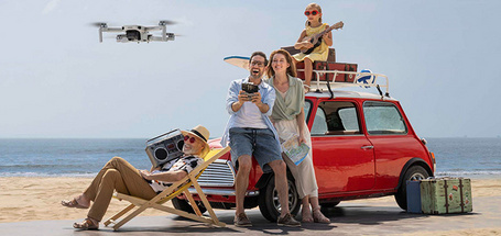 Famille à la plage avec une voiture rouge, un drone en vol, une chaise longue, des bagages et un enfant jouant du ukulélé.