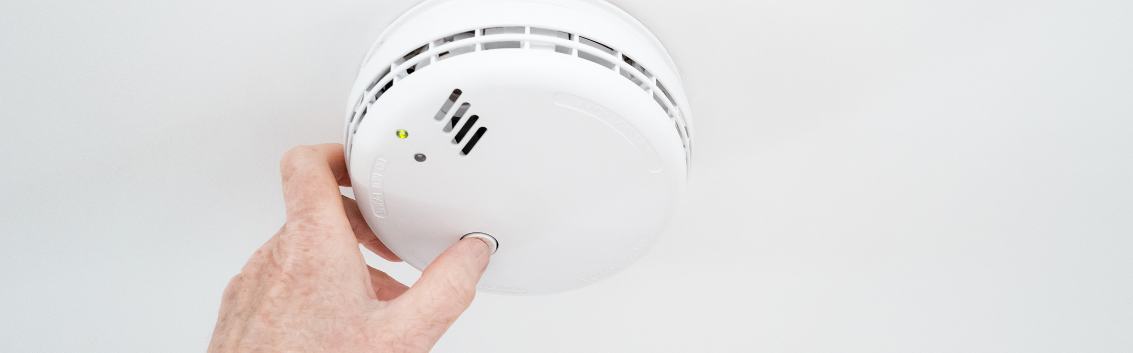 Une personne teste un détecteur de fumée fixé au plafond en appuyant sur le bouton de test.