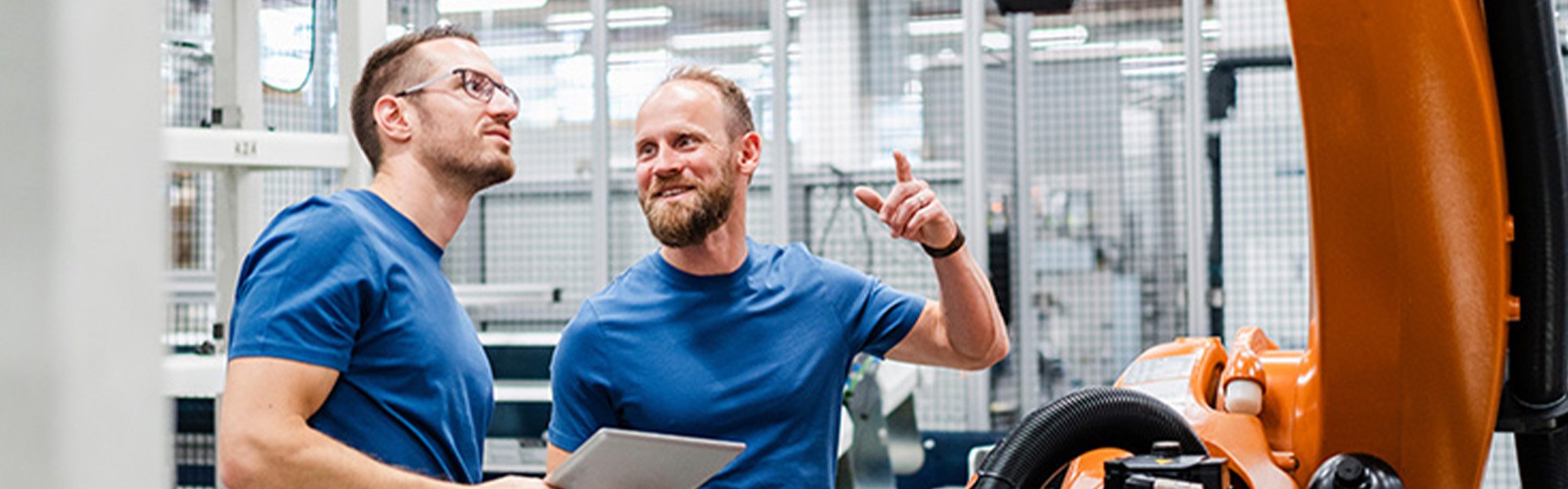 Deux hommes en t-shirt bleu parlent dans un hall d'usine devant un grand bras robotisé industriel.