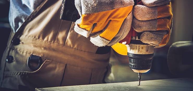Une personne portant des gants de travail visse une vis dans un matériau solide à l'aide d'une visseuse sans fil.